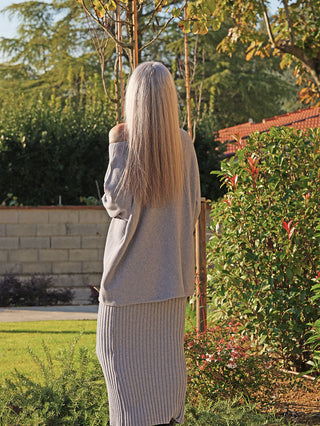 Person von hinten in einem Garten, trägt den grauen OFF#DLY Longpulli Rosi. Der Pulli hat einen lässigen, oversized Schnitt und off-shoulder Look, kombiniert mit einem gerippten Rock.