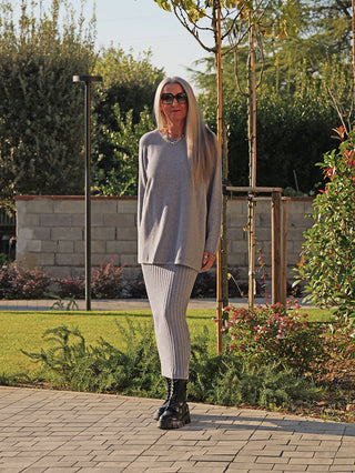 Ältere Frau trägt den OFF#DLY Longpulli Rosi grey, ein oversized, off-shoulder Strickset mit Pullover und geripptem Rock, abgerundet mit klobigen schwarzen Stiefeln, im Garten stehend.