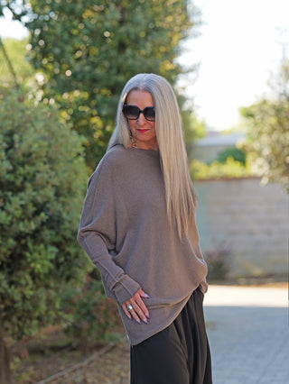 Frau trägt OFF#DLY Pulli Irene asymetric taupe mit Daumenlöchern. Der Pullover hat einen einzigartigen, asymmetrischen Schnitt, kombiniert mit weiten schwarzen Hosen und mehreren Ringen.