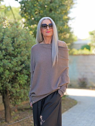 Eine Person trägt den OFF#DLY Pulli Irene asymetric taupe, einen locker geschnittenen, asymmetrischen Pullover mit offener Schulterpartie, kombiniert mit weit geschnittenen schwarzen Hosen, während sie draußen steht.