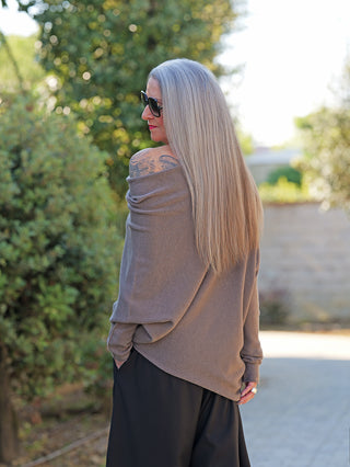 Frau in OFF#DLY Pulli Irene asymetric taupe, der locker über die Schulter fällt und ein Tattoo freilegt, kombiniert mit weiten schwarzen Hosen, während sie sich über die Schulter zur Kamera umdreht.