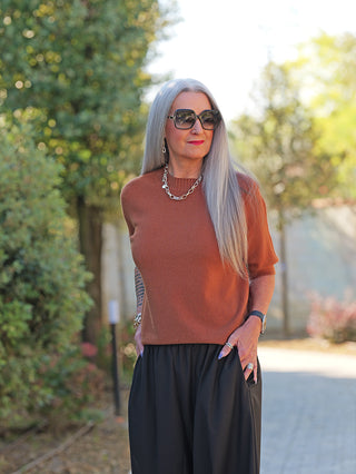 Frau trägt OFF#DLY Pulli Tina camel, kurzärmelig, figurbetont, kombiniert mit lockerer schwarzer Hose und Accessoires. Ideal für vielseitige Looks, zeigt modernes, schlichtes Design.