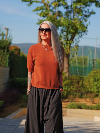 Frau trägt den OFF#DLY Pulli Tina camel, ein kurzärmliges, rust-oranges Oberteil mit schmalem Schnitt, kombiniert mit schwarzen Hosen und Accessoires. Ideal für einen modernen, vielseitigen Look.