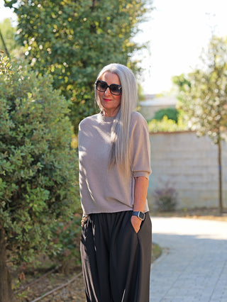 Frau trägt den OFF#DLY Pulli Tina beige, ein kurzärmeliges, schmal geschnittenes Oberteil, kombiniert mit schwarzen Hosen. Perfekt für einen modernen, vielseitigen Look in Büro oder Freizeit.