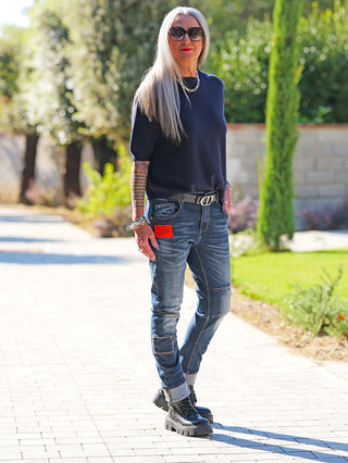 Person trägt OFF#DLY Pulli Tina darkblue, kurzärmelig und schmal geschnitten, kombiniert mit gepatchten Jeans und klobigen Stiefeln, in einem sonnigen Gartenweg.