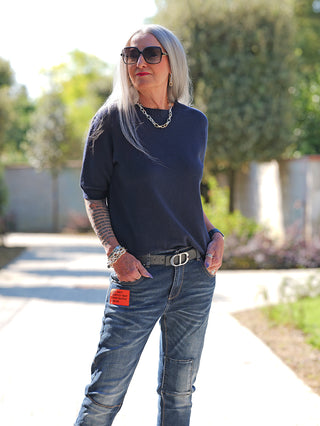 Ältere Frau trägt OFF#DLY Pulli Tina darkblue mit kurzen Ärmeln und schmalem Schnitt, kombiniert mit blauen Jeans. Sie steht auf einem sonnigen Weg, umgeben von einem unscharfen Gartenhintergrund.