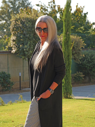 Frau trägt den OFF#DLY Strickmantel Laura black, ein lang geschnittener, offener schwarzer Cardigan mit Taschen, über einem schwarzen Oberteil, kombiniert mit hell gemusterten Hosen und einer auffälligen Halskette.