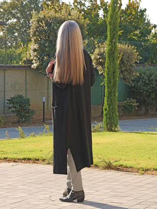 Person von hinten in einem langen schwarzen OFF#DLY Strickmantel Laura, kombiniert mit gemusterten Hosen und schwarzen Stiefeletten, stehend auf einem gepflasterten Weg vor einem grünen Gartenhintergrund.