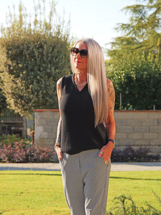 Frau in Garten trägt das OFF#DLY Stricktop Susi schwarz, ärmellos mit V-Ausschnitt. Kombiniert mit auffälligen Accessoires und gemusterten Hosen, zeigt sich der Stricktop vielseitig tragbar.