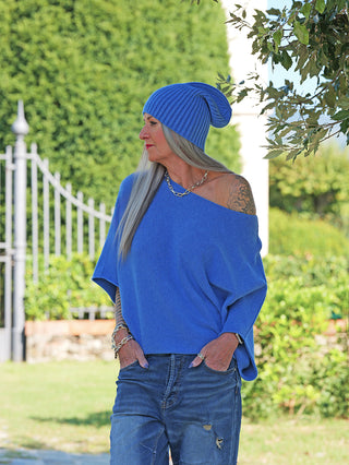 Person im Garten trägt den OFF#DLY Pulli Ellen denimblue, ein locker geschnittenes, schulterfreies Oberteil mit Fledermausärmeln. Kombiniert mit Jeans, silbernem Schmuck und einer blauen Strickmütze.