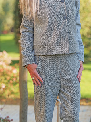 Person trägt OFF#DLY Pant Lina black/white Anzughose in grauem Tweed mit feinem Muster. Hose hat schmalen Schnitt, elastischen Bund. Hände teils in Taschen, umgeben von Gartenkulisse.