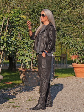 Frau trägt OFF#DLY Pant Lani black in Lederoptik mit elastischem Bund, kombiniert mit passender Jacke, während sie eine Zitrusfrucht inspiziert. Ihre Accessoires: Smartwatch, Armbänder, Tattoo.