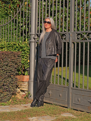 Erwachsener lehnt lässig an einem Tor, trägt OFF#DLY Pant Lani in schwarzer Lederoptik mit lockerem Schnitt und elastischem Bund, kombiniert mit schwarzer Jacke und Accessoires.