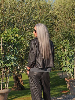 Eine silberhaarige Frau trägt die OFF#DLY Jacke Luisa black mit modischem, kurzem Schnitt und stilvoller Lederoptik in einem sonnenbeschienenen Garten, passend kombiniert zu dunklen Hosen.