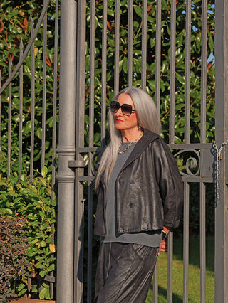 Die OFF#DLY Jacke Luisa black zeigt eine elegante Lederoptik mit modernem, kurzem Schnitt und Knopfleiste, getragen von einer Frau vor einem dekorativen Tor im Freien.