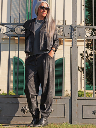 Die OFF#DLY Jacke Luisa black präsentiert sich in moderner Lederoptik mit kurzem, kastigem Schnitt und Knopfleiste, getragen von einer stilvollen Frau vor einem schmiedeeisernen Tor.