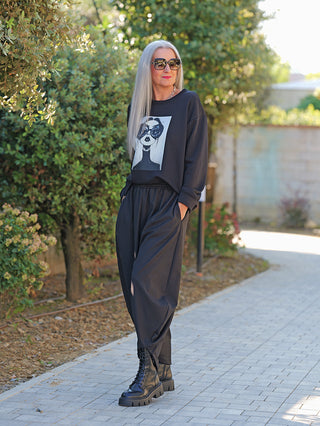 Frau mit langen, grauen Haaren trägt OFF#DLY Sweat Ela black, ein schwarzes Oversize-Sweatshirt mit stilisiertem Gesichtsdruck, kombiniert mit weiten Hosen und klobigen Stiefeln auf einem Gartenweg spazierend.