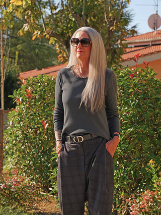 Erwachsene Person im Garten trägt das OFF#DLY LS Alice antra Longsleeve, schmal geschnitten, mit Rippstruktur und offenen Kanten, kombiniert mit Sonnenbrille und Schmuck, vor grünen Sträuchern.