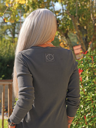 Die Frau trägt das stilvolle OFF#DLY LS Alice antra Langarmshirt mit schmalem Schnitt und Rippstruktur, verziert mit einem dezenten Label-Patch auf der Rückseite.