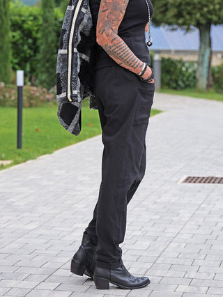 Person trägt OFF#DLY Pant Bora black mit lockerem Schnitt und Seitentasche; kombiniert mit schwarzem Top, Lederboots und über Schulter geworfenem Pullover. Floral tätowierter Arm sichtbar.