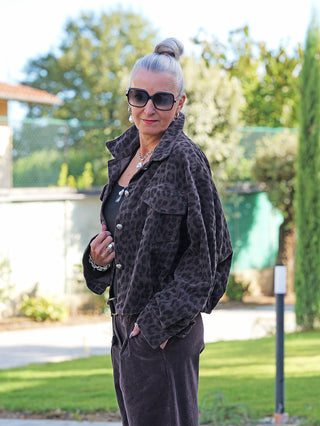 Ältere Frau im Garten trägt die OFF#DLY Cordjacke Valerie schoko/leo, mit lässigem Leopardenmuster, über einem schwarzen Top, kombiniert mit dunklen Hosen und silbernem Schmuck.