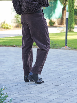Person in OFF#DLY Pant Vada schoko aus dunklem Cord, steht auf einem gepflasterten Bereich. Die Hose hat einen lockeren Schnitt und elastischen Bund, kombiniert mit schwarzen Stiefeletten.