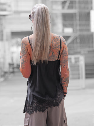 Person mit Rückenansicht trägt das OFF#DLY Top Violetta schwarz, ein schwarzes Camisole-Top mit Spitze am Saum, passend zu neutralen Cargo-Hosen. Tätowierte Arme und Rücken.