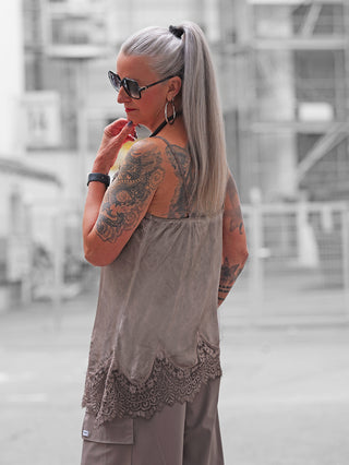 Frau mit silbernem Haar trägt das OFF#DLY Top Violetta in Taupe, ein locker fallendes, spitzenbesetztes Top, kombiniert mit weiten Hosen, urbaner Hintergrund mit Baustellenelementen.