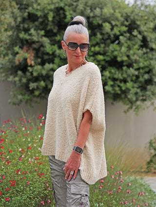 Person im Garten trägt den OFF#DLY Pullunder Stella creme, ein locker geschnittenes, grob gestricktes Top mit V-Ausschnitt, kombiniert mit grauen Cargo-Hosen und Accessoires. Ideal für Layering und lässigen Stil.