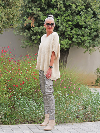 Person trägt den OFF#DLY Pullunder Stella creme, einen locker geschnittenen, grob gestrickten Pullunder mit V-Ausschnitt, über grauen Cargo-Hosen und beigefarbenen Stiefeletten, ergänzt durch auffällige Accessoires.