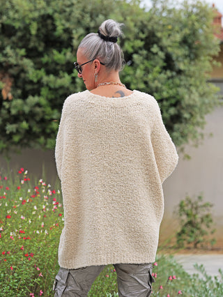 Person von hinten in Garten, trägt OFF#DLY Pullunder Stella creme: oversized, grob gestrickt, V-Ausschnitt. Silbriges Haar im Dutt, Sonnenbrille, Kettenhalsband. Kombiniert mit olivgrüner Cargo-Hose.