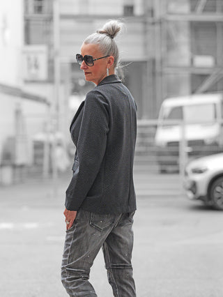 Person trägt den OFF#DLY Blazer Olga grau mit klassischem Reverskragen und taillierter Passform. Kombiniert mit Jeans, vermittelt der Blazer eine elegante, zeitlose Silhouette.