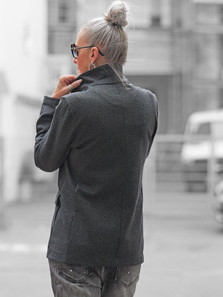 Person von hinten in schmaler Gasse, trägt OFF#DLY Blazer Olga grey mit leicht taillierter Passform, klassischem Reverskragen, hebt den Kragen an, kombiniert mit grauen Jeans und silbernen Ohrringen.