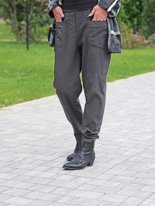 Person trägt OFF#DLY Pant Bora grau, locker geschnitten mit großen Fronttaschen und elastischem Bund, kombiniert mit schwarzen Stiefeletten. Ideal für urbane Looks mit modernem, bequemen Stil.