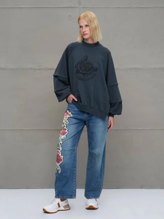 Junge Person im #VDR Sweatshirt 18156 anthrazit mit übergroßem Schnitt, Rundhalsausschnitt und großer Rosen-Grafik, kombiniert mit Jeans und Sneakers, vor grauer Wand stehend.