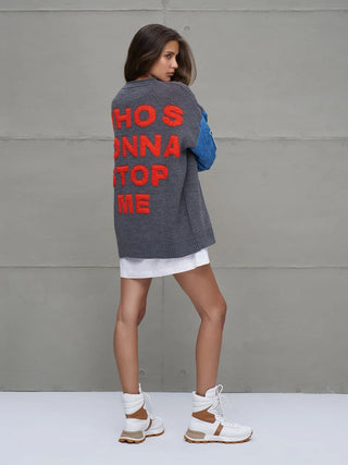 Junge Frau trägt #VDR Knit Sweat 18073 grey mel, einen übergroßen Strickpullover mit auffälligem Rückenschriftzug „WHO'S GONNA STOP ME“, kombiniert mit kurzem weißen Rock und weißen Stiefeletten.