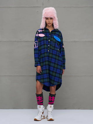 Junge Frau trägt #VDR Shirt 18114 royal/black, ein übergroßes Plaid-Kleid mit Applikationen, kombiniert mit pinkem Trapper-Hut, grauen Socken und weißen Stiefeln, vor grauer Wand.