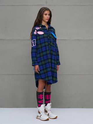 Junge Frau trägt #VDR Shirt 18114 royal/black, ein oversized Hemdkleid mit auffälligem Karomuster und bunten Patches, kombiniert mit kniehohen Socken und weißen Stiefeln.