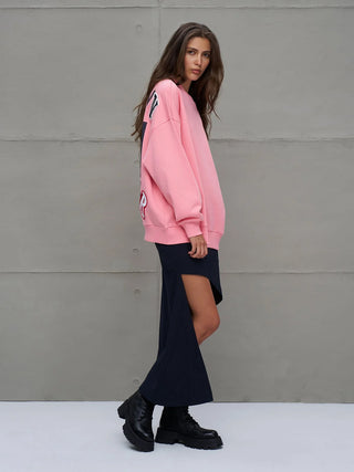 Junge Frau in #VDR Sweater 18218 pink, oversized mit Rundhalsausschnitt, Statement-Slogan auf der Rückseite, kombiniert mit langem Rock und schwarzen Stiefeln, präsentiert einen lässigen Streetstyle-Look.
