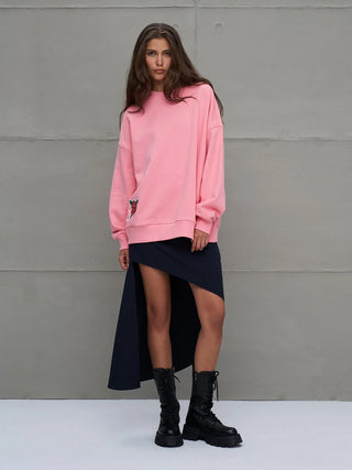 Eine junge Frau trägt den #VDR Sweater 18218 pink, ein oversized Sweatshirt mit aufgesticktem Patch. Ihr Outfit kombiniert einen asymmetrischen Rock und schwarze Stiefel, während sie direkt in die Kamera schaut.