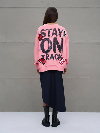 #VDR Sweater 18218 pink: Frau von hinten in einem oversized, bubble-gum-pinken Sweatshirt mit STAY ON TRACK Print und Patches, kombiniert mit dunklem Rock und schwarzen Boots, vor Betonwand stehend.