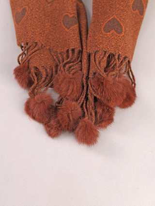 Der #OFF#DLY Schal Heart braun präsentiert sich in einem rust-orange Strick mit Herzmuster und langen Fransen, die mit flauschigen Pom-Pom-Quasten verziert sind.