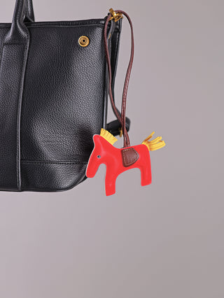 Nahaufnahme der Enrico Coveri Tote Bag black aus schwarzem genarbtem Leder mit rotem, pferdeförmigem Anhänger am Henkel. Der Anhänger hat eine braune Satteldekoration und gelbe Fransen.