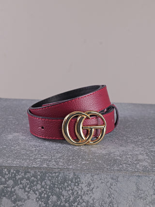 Der OFF#DLY Ledergürtel CG bordeaux zeigt sich in rotem Leder mit einer glänzenden, goldfarbenen Schnalle aus zwei ineinandergreifenden Ringen, kunstvoll auf einer steinähnlichen Fläche arrangiert.