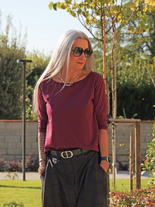 Frau im Garten trägt das OFF#DLY LS Lahela bordeaux, ein locker geschnittenes, elegantes Langarmshirt, kombiniert mit dunklen Karohosen und Accessoires wie einer Kette und Sonnenbrille.
