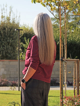 Person im Garten mit dem schmal geschnittenen OFF#DLY LS Lahela bordeaux Langarmshirt, das die Figur betont, kombiniert mit einer Smartwatch am Handgelenk und dunkler Hose, von hinten fotografiert.