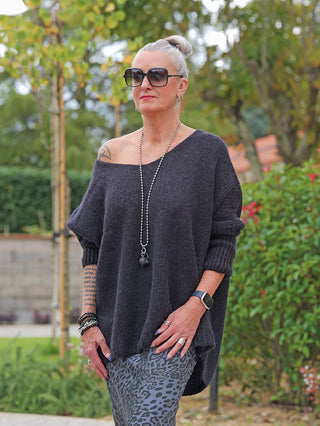 Frau im Garten trägt OFF#DLY Pulli Selma black, lässig-weite Passform, off-shoulder, mit Mohairanteil. Kombiniert mit grauen Leggings, Accessoires und Tattoos sichtbar.