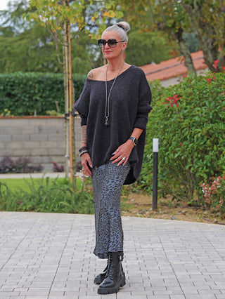 Frau trägt den OFF#DLY Pulli Selma black, einen lässig-geschnittenen, schulterfreien Pullover aus weichem Strick, kombiniert mit einem langen Leopardenrock und robusten Stiefeln, Accessoires inklusive.
