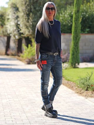 Frau trägt OFF#DLY Denim Life Dark Blue mit Patches und rotem Etikett. Kombiniert mit schwarzem Gürtel, Kettenhalskette und klobigen Stiefeln. Perfekter urbaner Look, lässig und komfortabel.