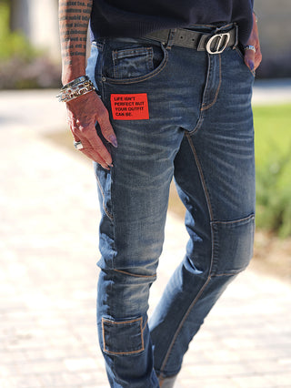 Nahaufnahme von OFF#DLY Denim Life Dark Blue Jeans mit Patchwork-Kniedetails und rotem Label, getragen mit schwarzem Gürtel und Silberschmuck, betont urbanen Look und Tragekomfort.